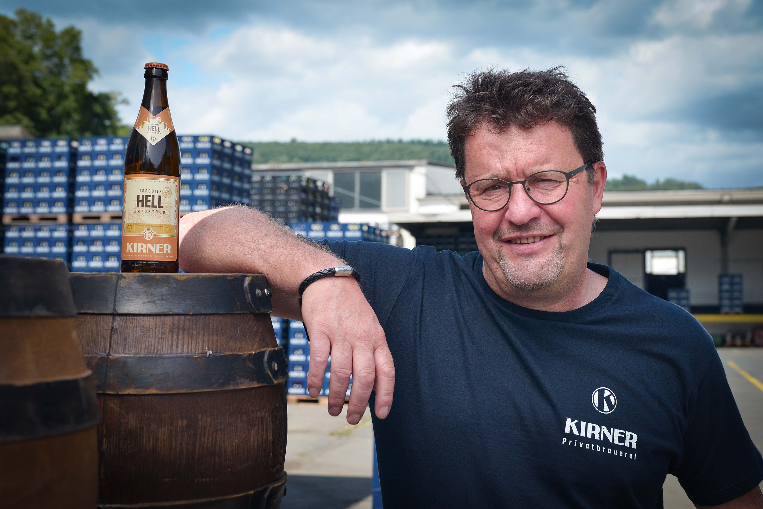 Kirner Mitarbeiter lehnt mit Ellenbogen auf Bierfass - Schaut lachend in die Kamera - Landbier steht auf Bierfaß